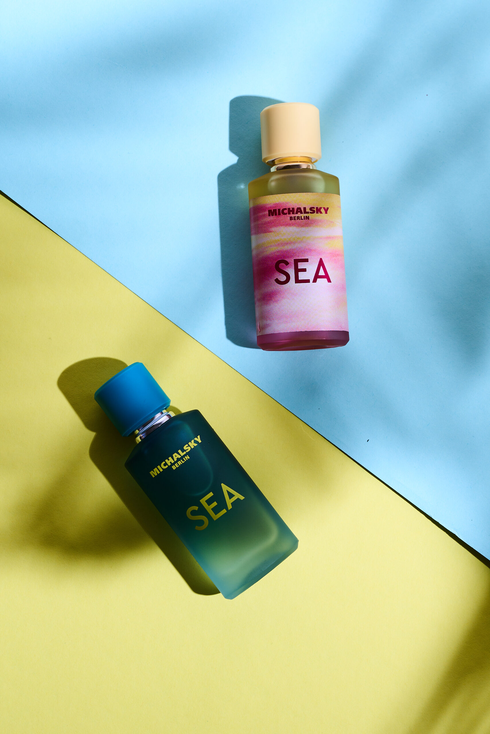 Zwei Michalsky Berlin SEA Parfumflakons in Blau und Rosa auf hellblauem und gelbem Hintergrund mit markanten Lichtschatten.